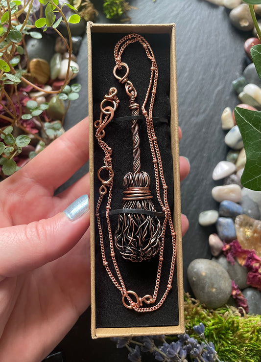 Copper necklace with broom pendant in a box held by a hand with a natural background of stones and plants.