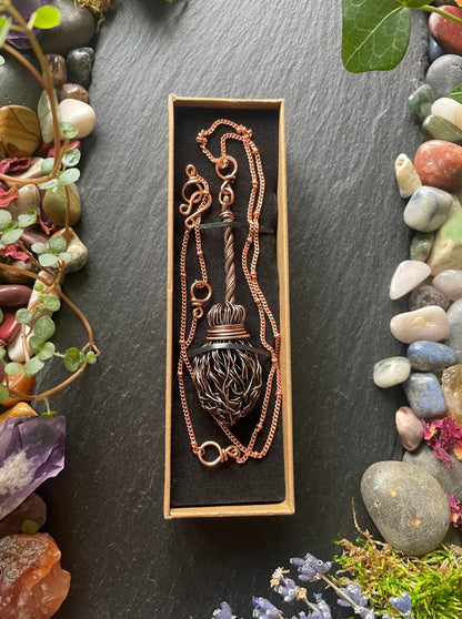 Copper broom necklace in a box on a dark stone surface with natural elements around