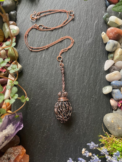 Decorative broom pendant on a dark surface with stones and plants.