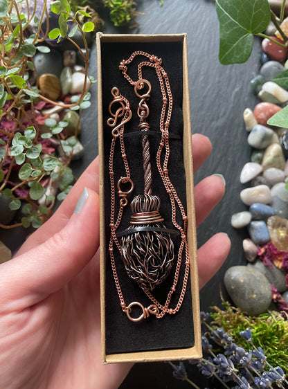 Copper broom necklace in a box held by a hand with a natural background of stones and plants.