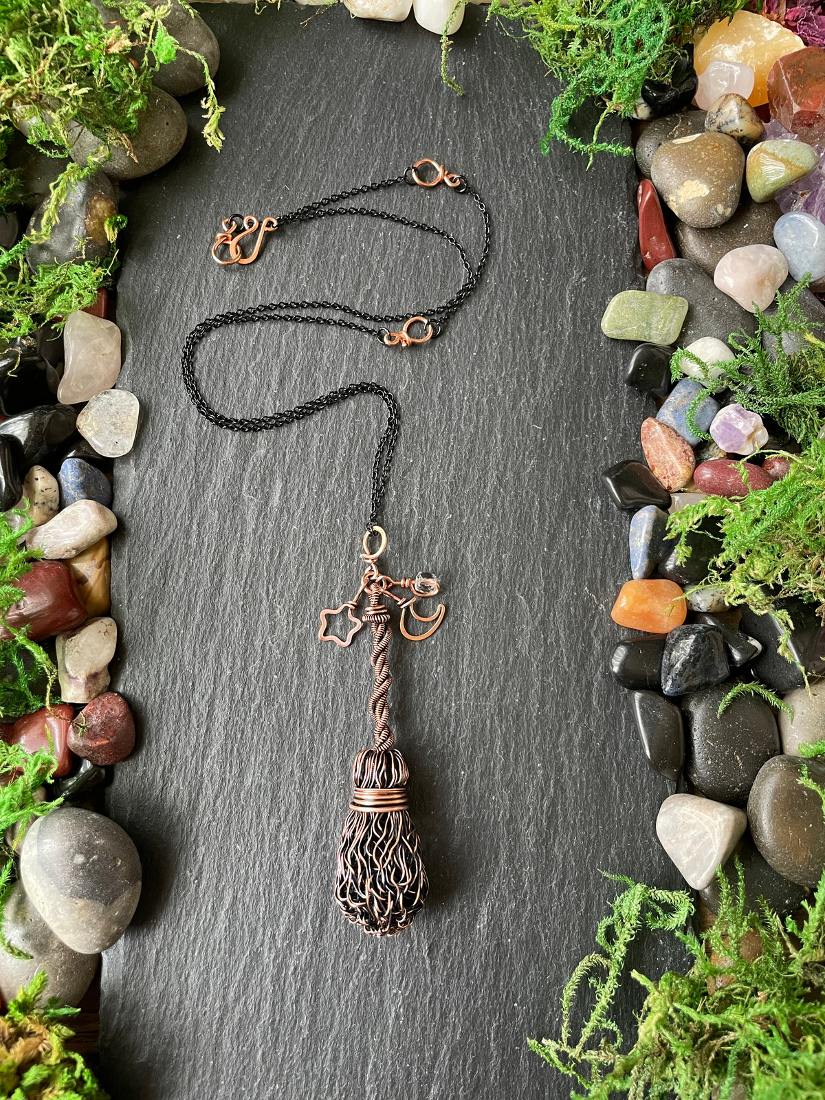 A broom pendant necklace, displayed on a dark slate background surrounded by moss and stones.