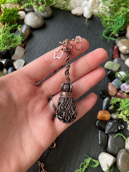 A hand holding a copper broom pendant, set against a background of moss and smooth stones.