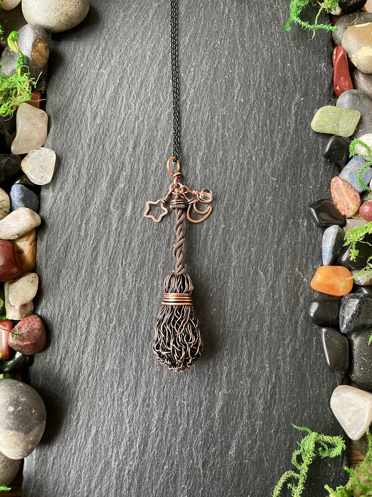 A handmade copper and brown wire pendant necklace shaped like a broom, hanging on a dark slate background framed by decorative stones and moss.