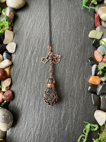 A handmade copper and brown wire pendant necklace shaped like a broom, hanging on a dark slate background framed by decorative stones and moss.
