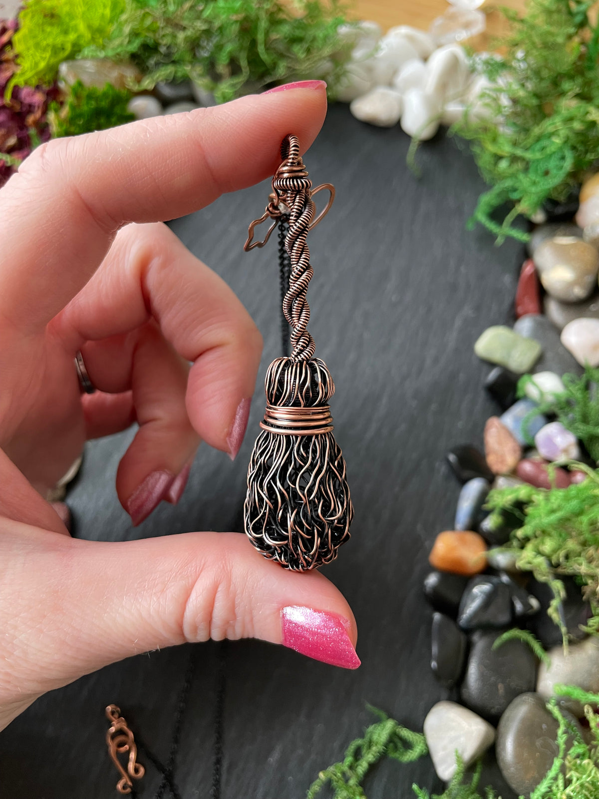 A hand holding a copper broom pendant, set against a dark slate background with decorative stones and greenery.