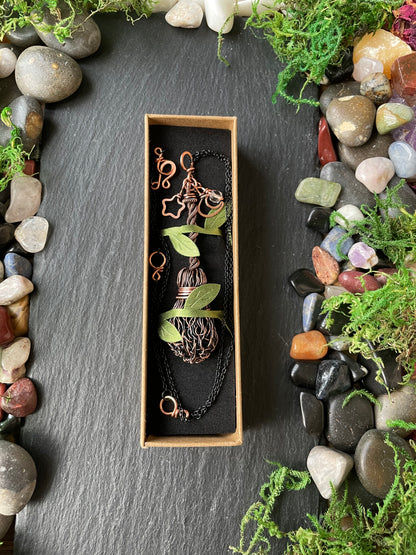 A copper broom pendant, displayed on a dark slate background surrounded by stones and moss.