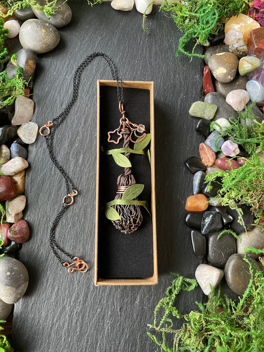 A handcrafted copper broom pendant on a stone slate; the scene is styled with smooth stones and green moss on a dark slate surface.