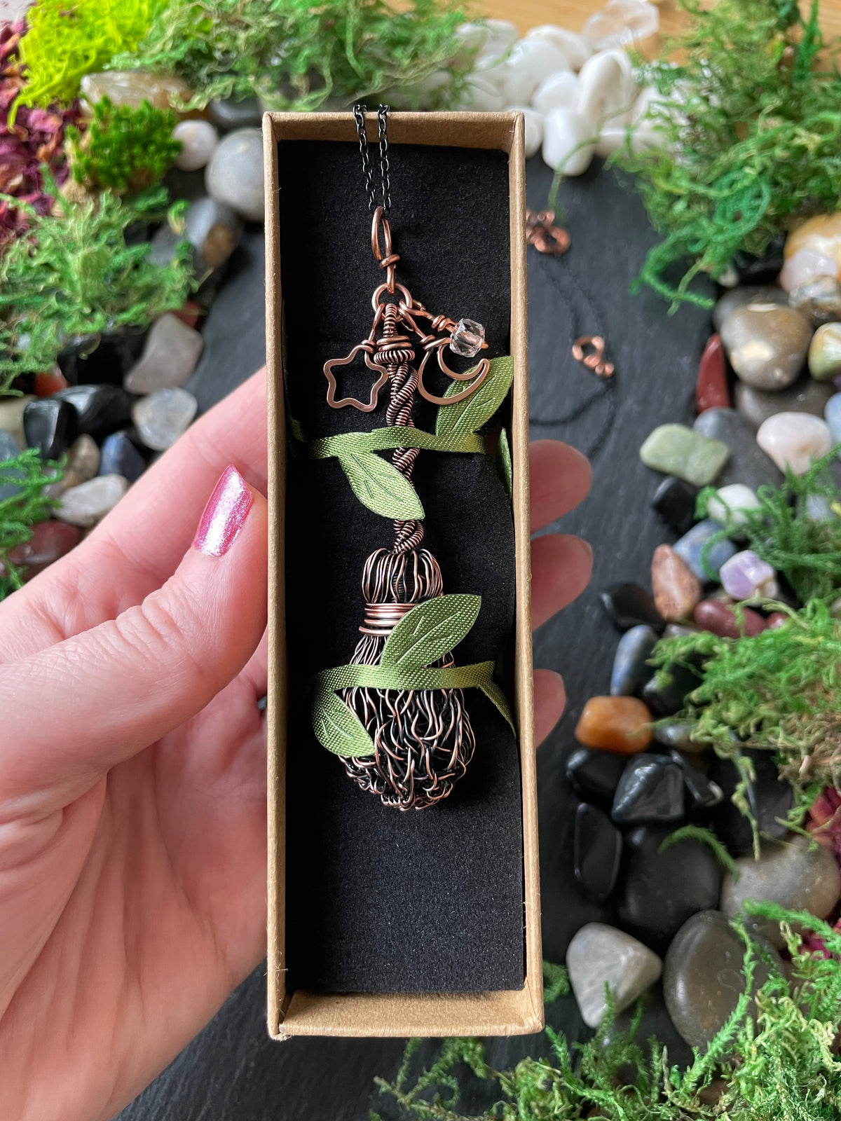 A hand holds a small rectangular jewelry box containing a copper-colored wire-wrapped pendant, set against a background of moss, stones, and small plants.
