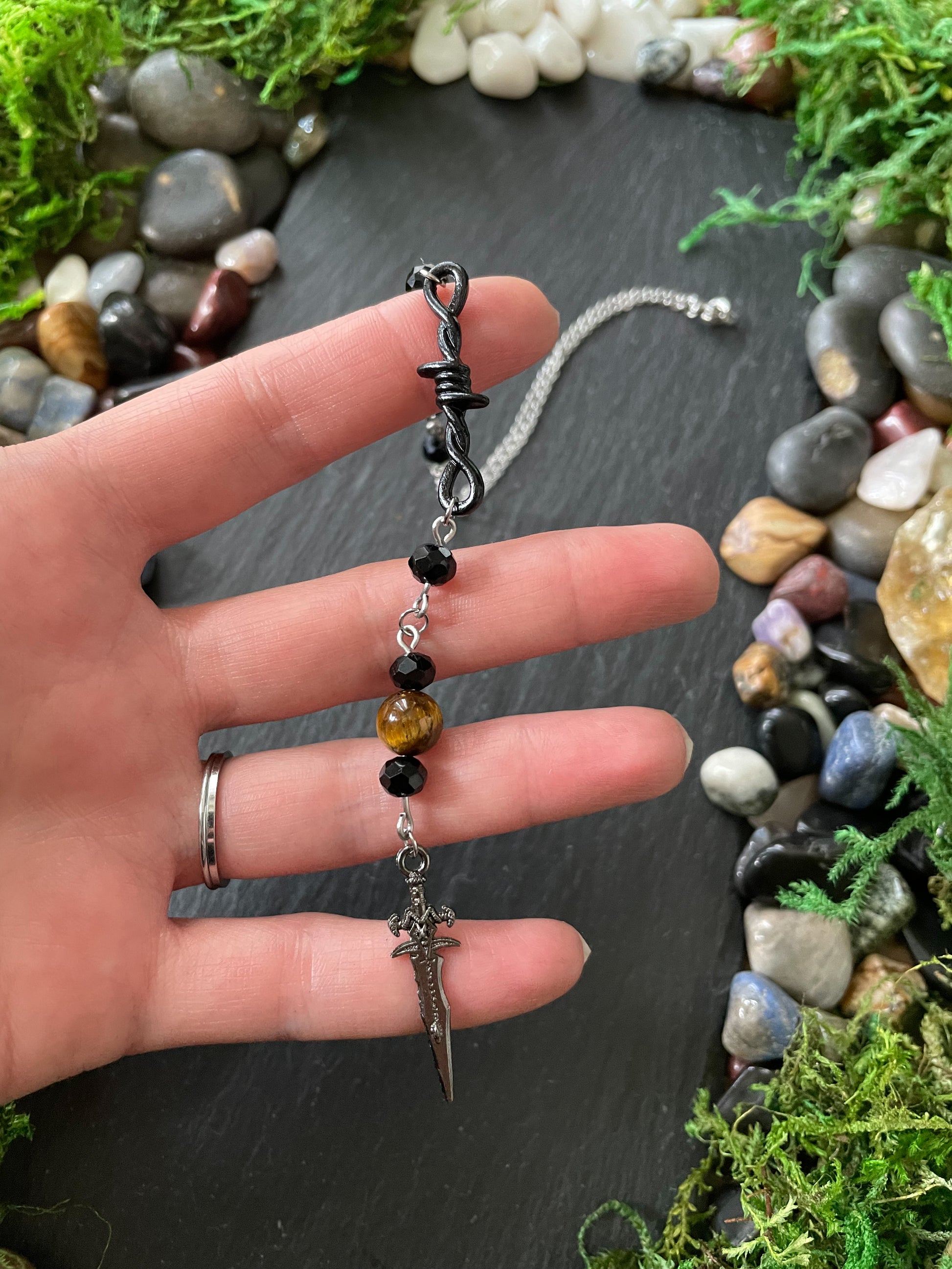 A hand holding a car charm with black beads, tiger's eye bead, and a silver dagger pendant, set against a dark slate background with moss and stones.