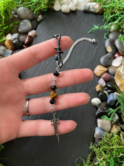 A hand holding a car charm with black beads, tiger's eye bead, and a silver dagger pendant, set against a dark slate background with moss and stones.