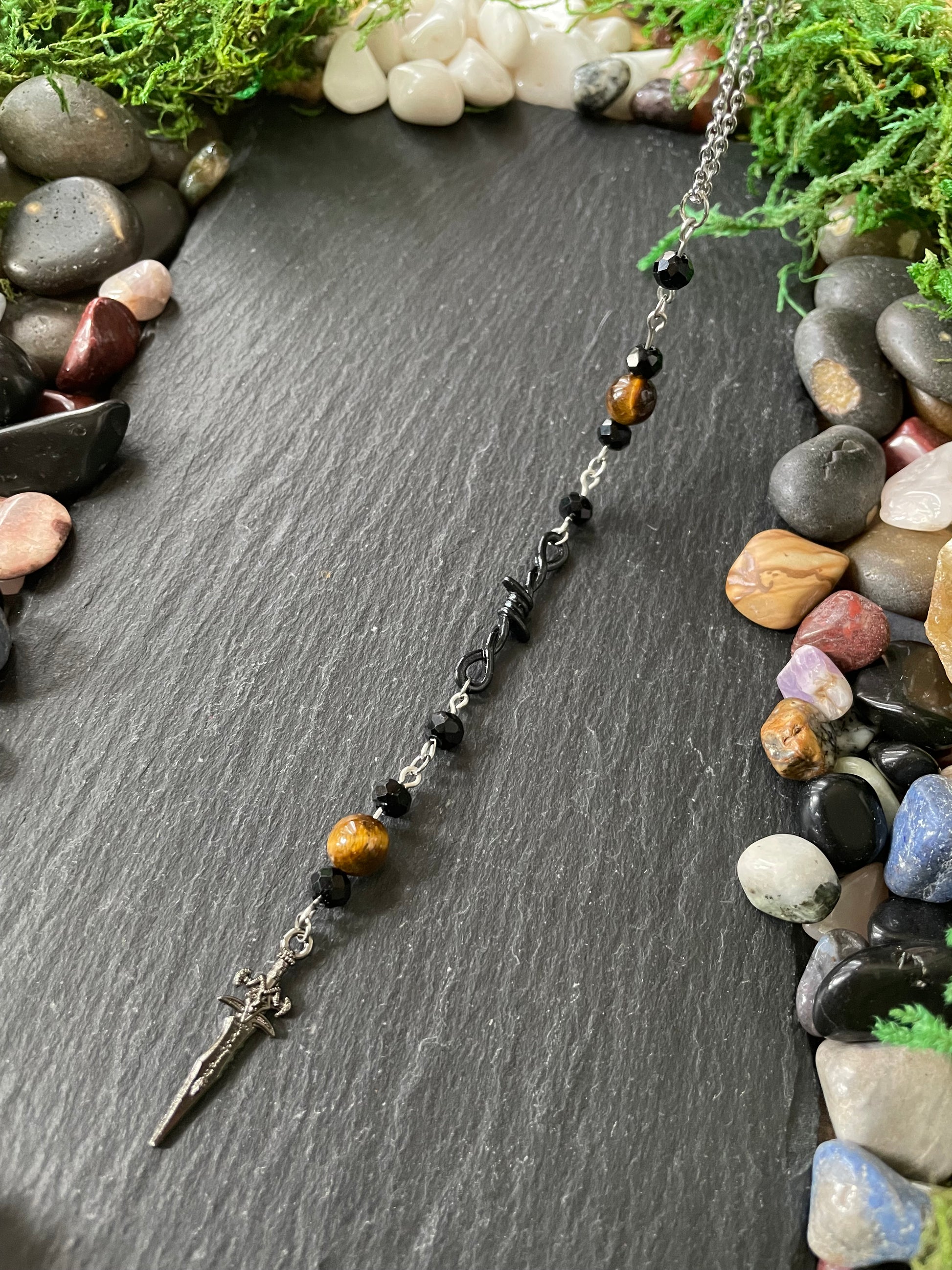 A beaded car charm featuring black beads, brown tiger's eye beads, and a silver dagger pendant, displayed on a dark slate background with decorative stones and moss.