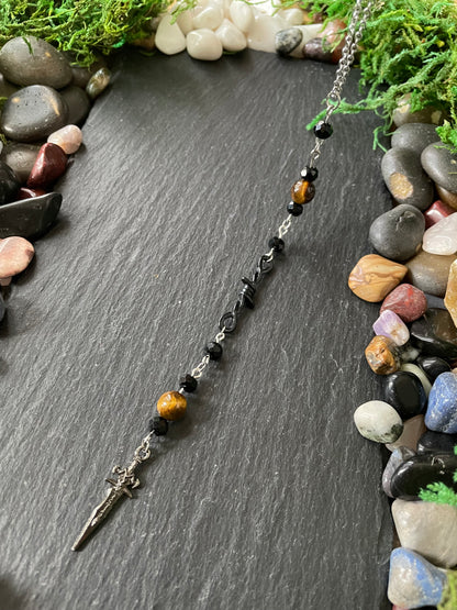 A beaded car charm featuring black beads, brown tiger's eye beads, and a silver dagger pendant, displayed on a dark slate background with decorative stones and moss.