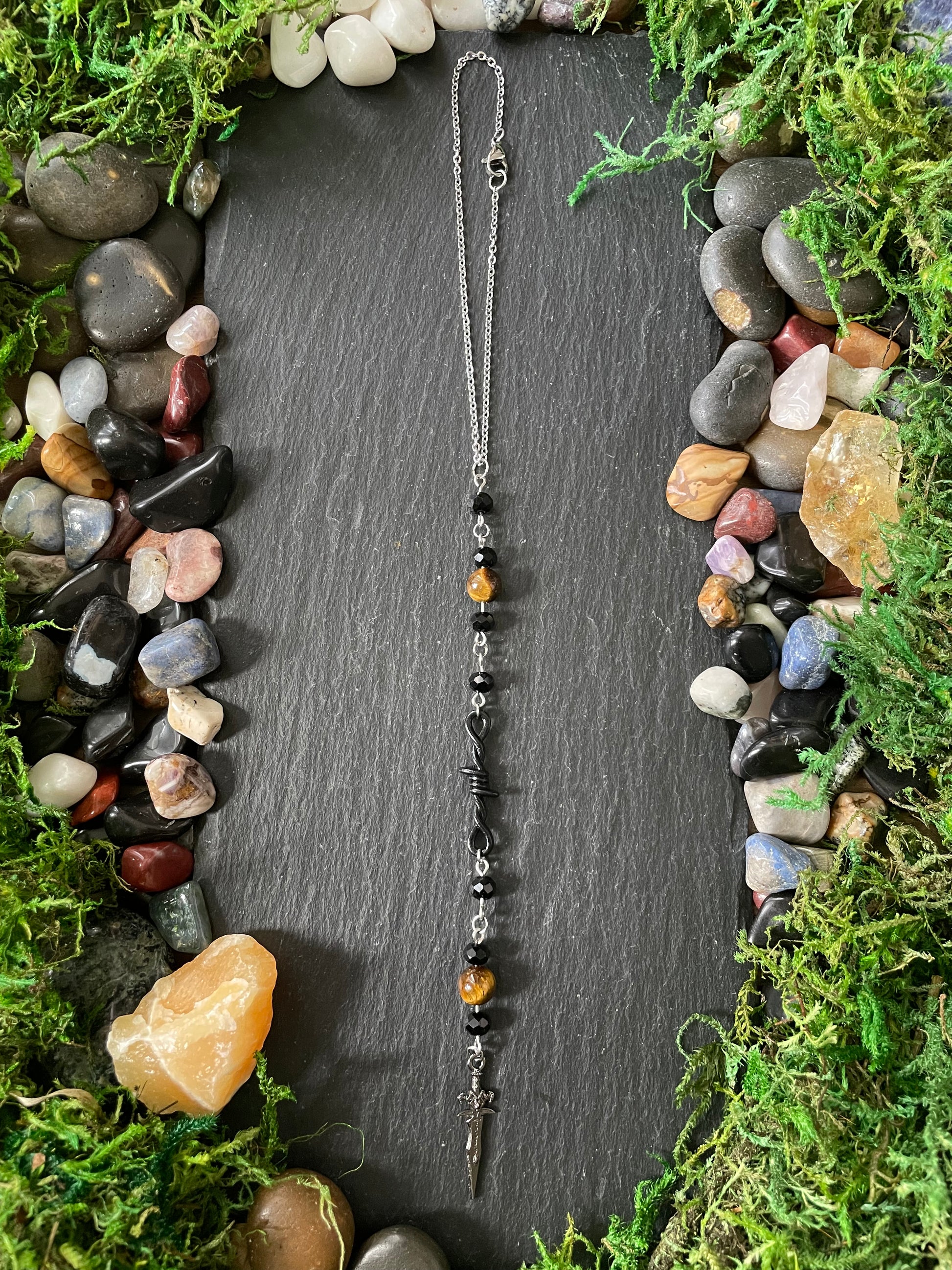 A car charm with amber and black beads, laid flat on a dark slate board surrounded by moss and small stones.