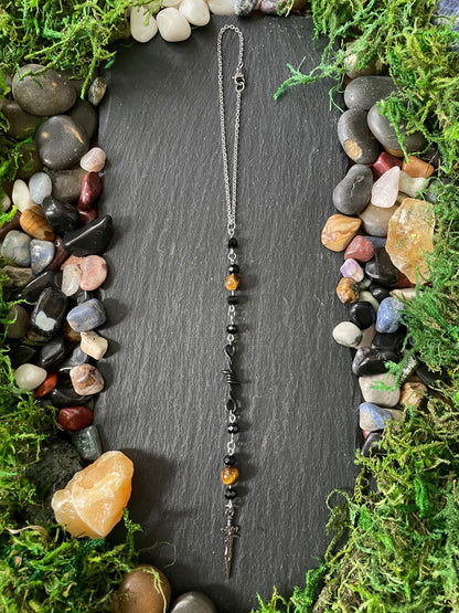 A car charm with amber and black beads, laid flat on a dark slate board surrounded by moss and small stones.