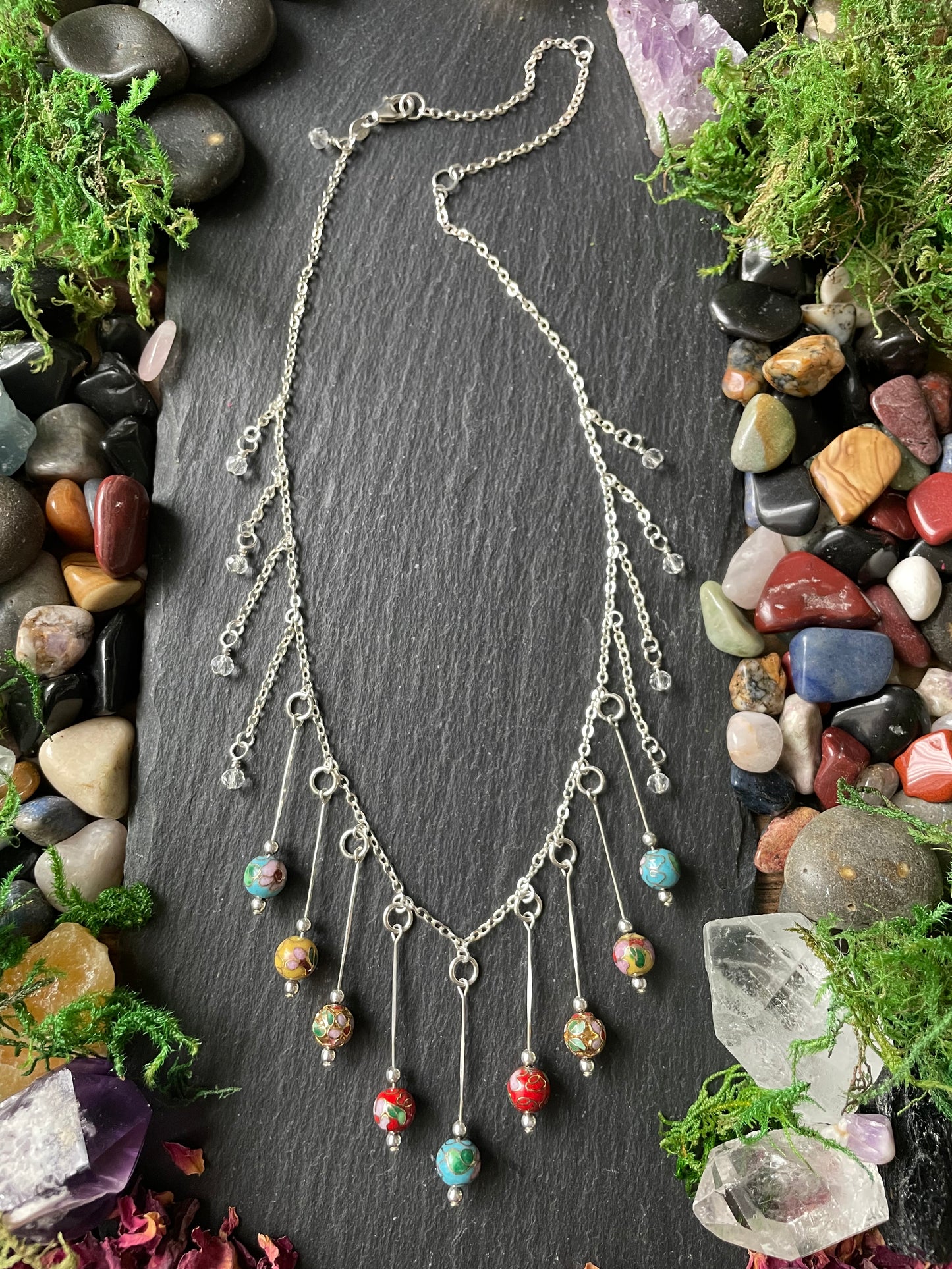 Silver necklace displayed on a dark stone surface with colorful stones and greenery around.