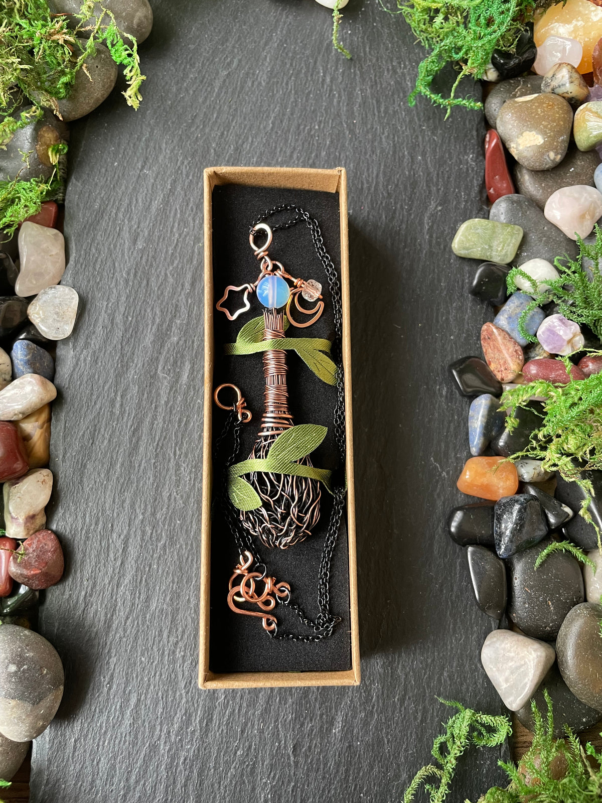 A copper-colored metal pendant necklace, presented in a small rectangular box on a dark slate background decorated with stones and greenery.