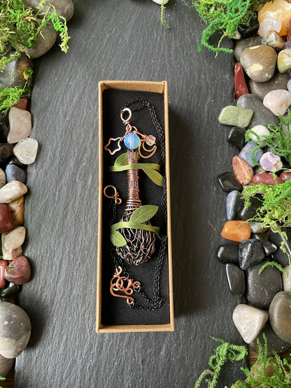 A copper-colored metal pendant necklace, presented in a small rectangular box on a dark slate background decorated with stones and greenery.