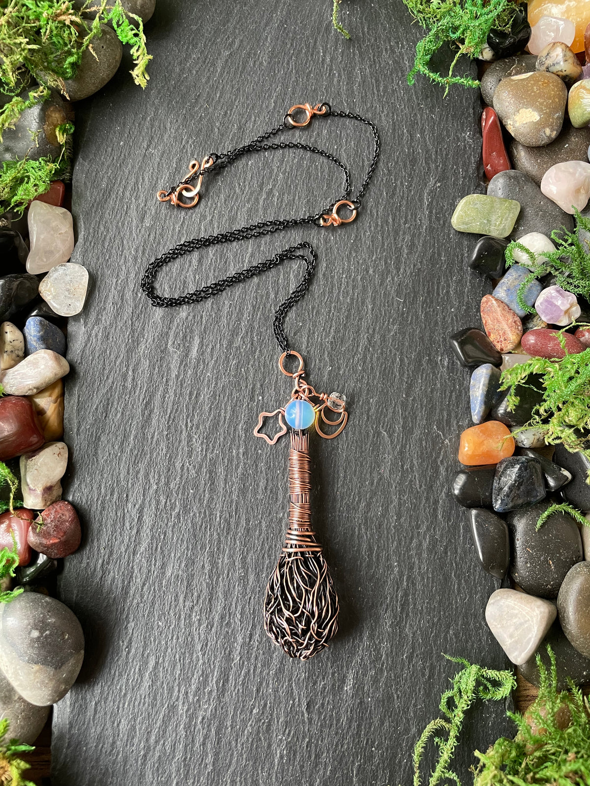 A handmade pendant necklace, displayed on a dark slate background surrounded by decorative stones and moss.