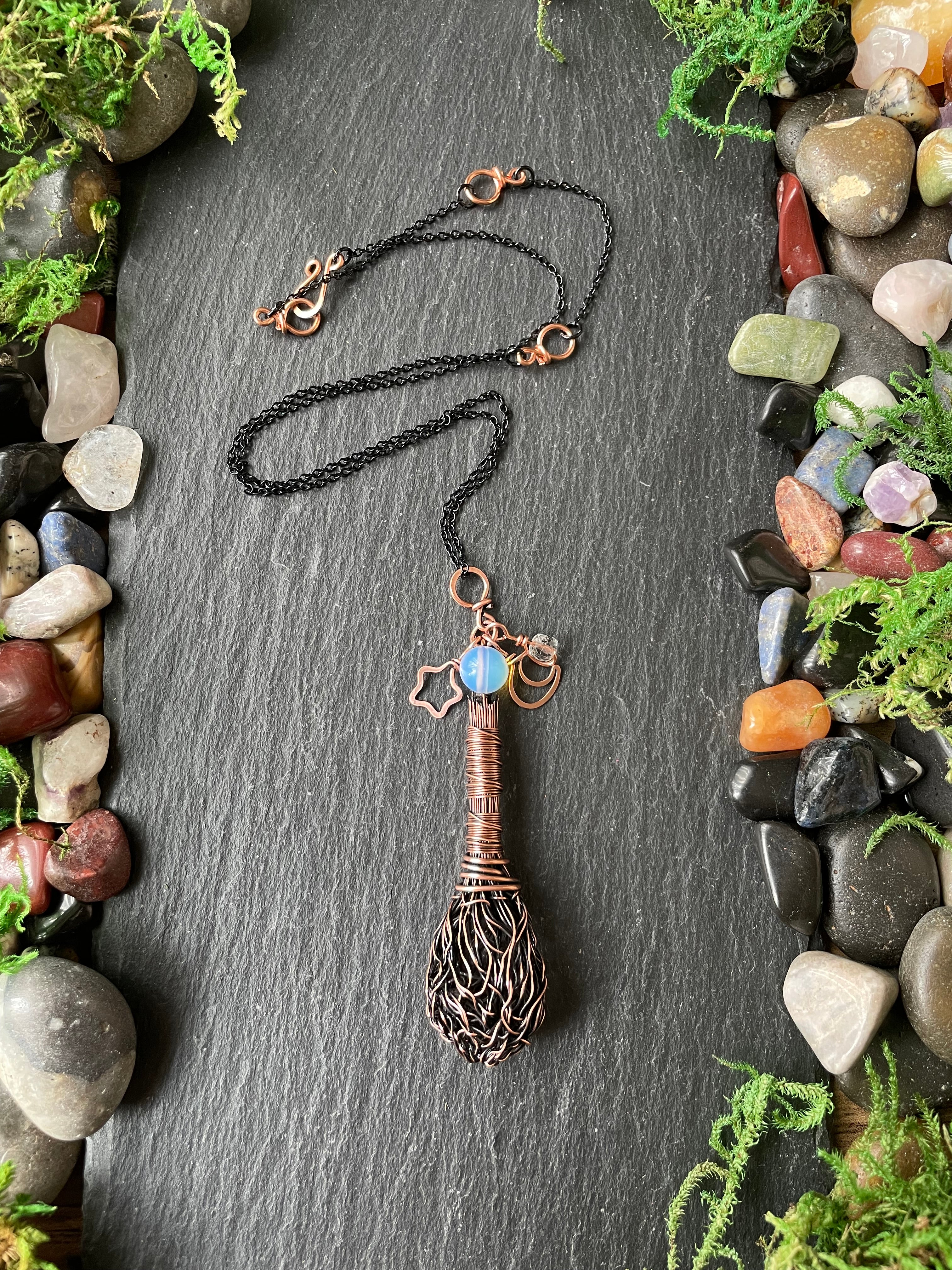 A handmade pendant necklace, displayed on a dark slate background surrounded by decorative stones and moss.