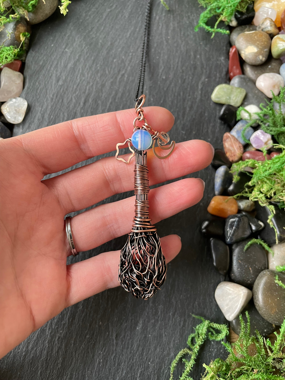 A hand holding a copper broom pendant, set against a dark slate background with decorative stones and moss.