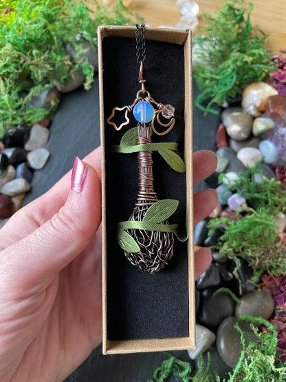 A hand hold a mini handmade broom pendant set against a background of moss and stones.