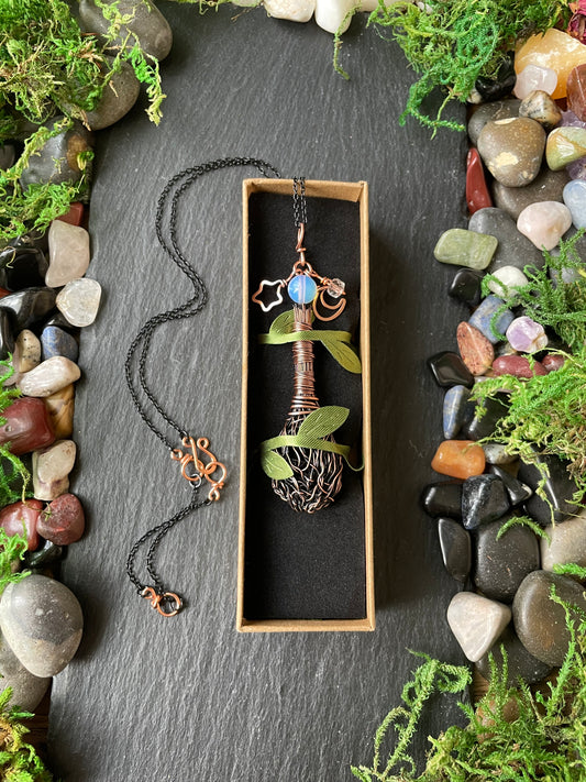 A handmade broom pendant, displayed in a wooden box on a black background with decorative stones and moss.