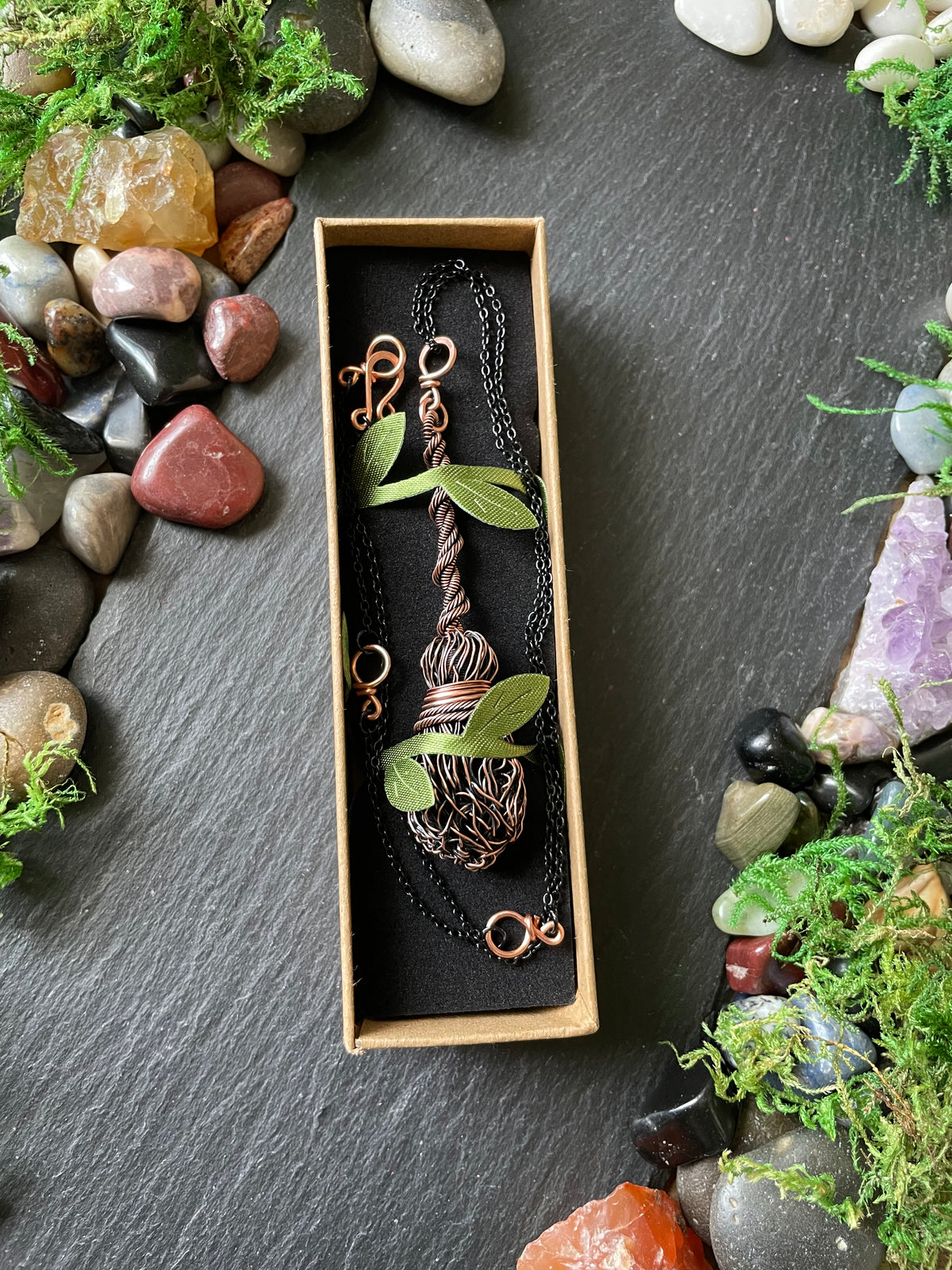 A broom pendant displayed in a small rectangular box on a black background. 