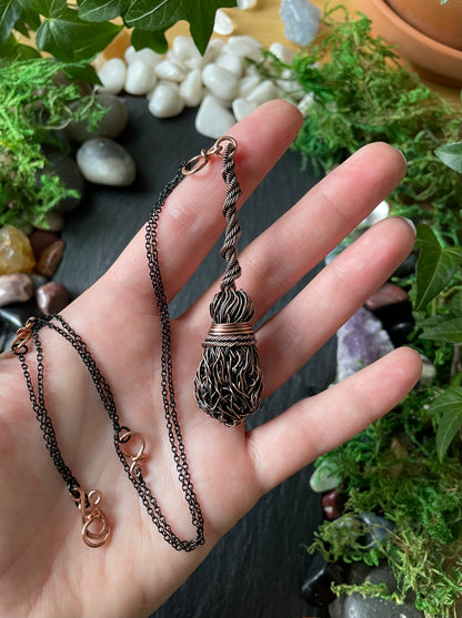 A dark copper pendant necklace with a textured, held in a person's hand against a background of green plants and small stones.