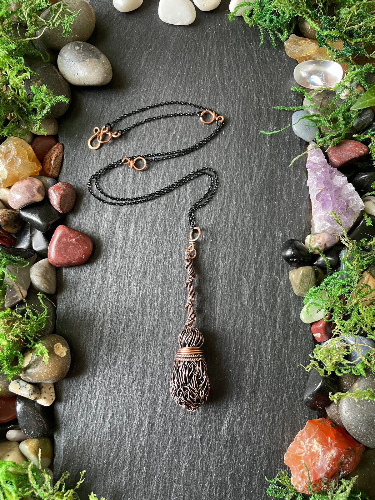 A handmade pendant necklace displayed on a dark slate background surrounded by decorative stones and moss.