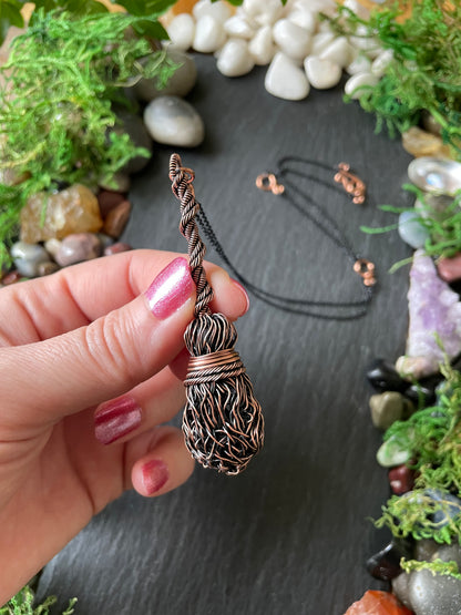 A hand holds a copper-colored broom pendant with a textured, wrapped head and a thin black chain. The background is a dark slate surface decorated with green moss.
