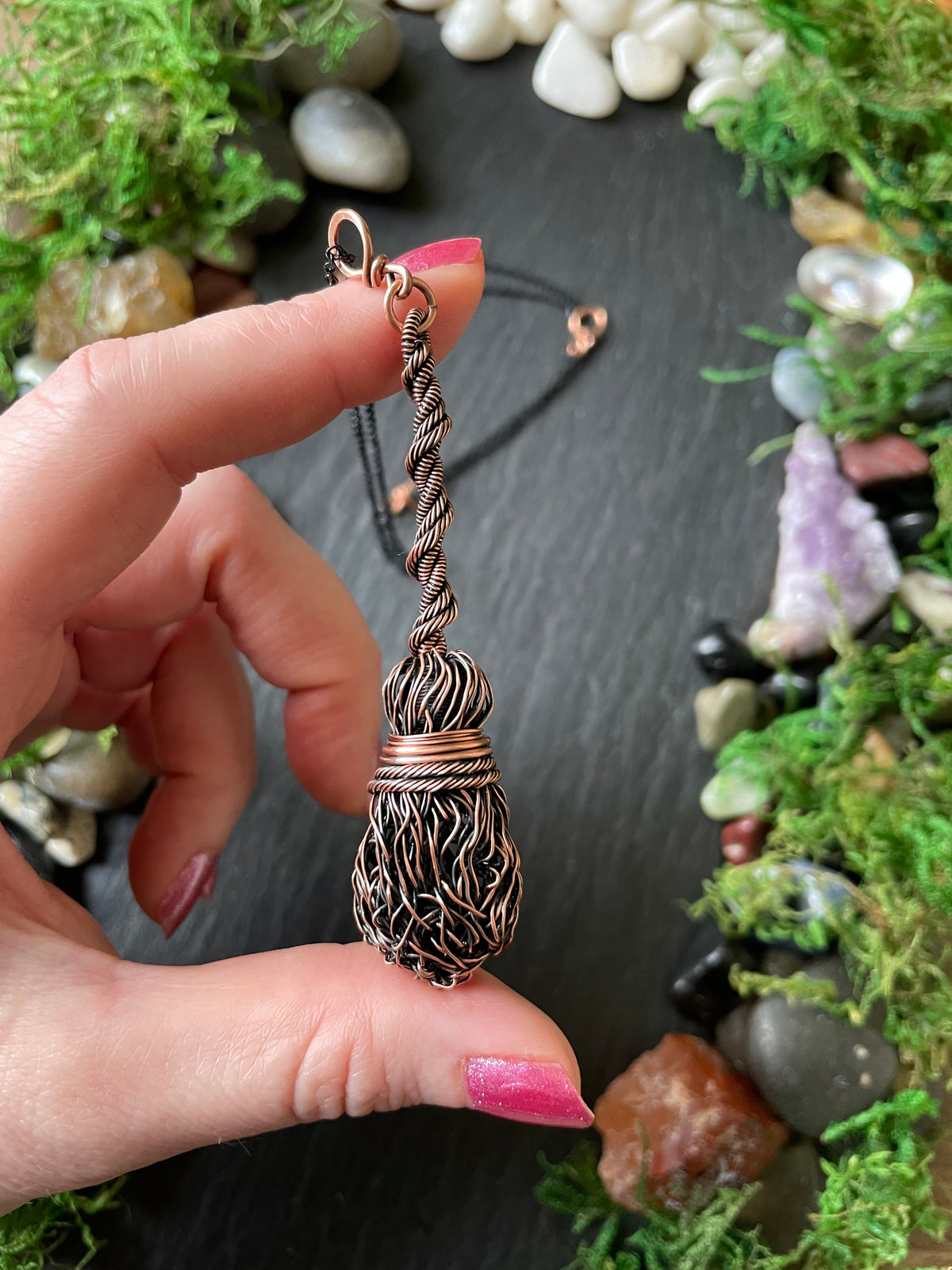 A hand holds a handmade copper wire pendant with a twisted handle, set against a dark slate background decorated with moss and small stones.