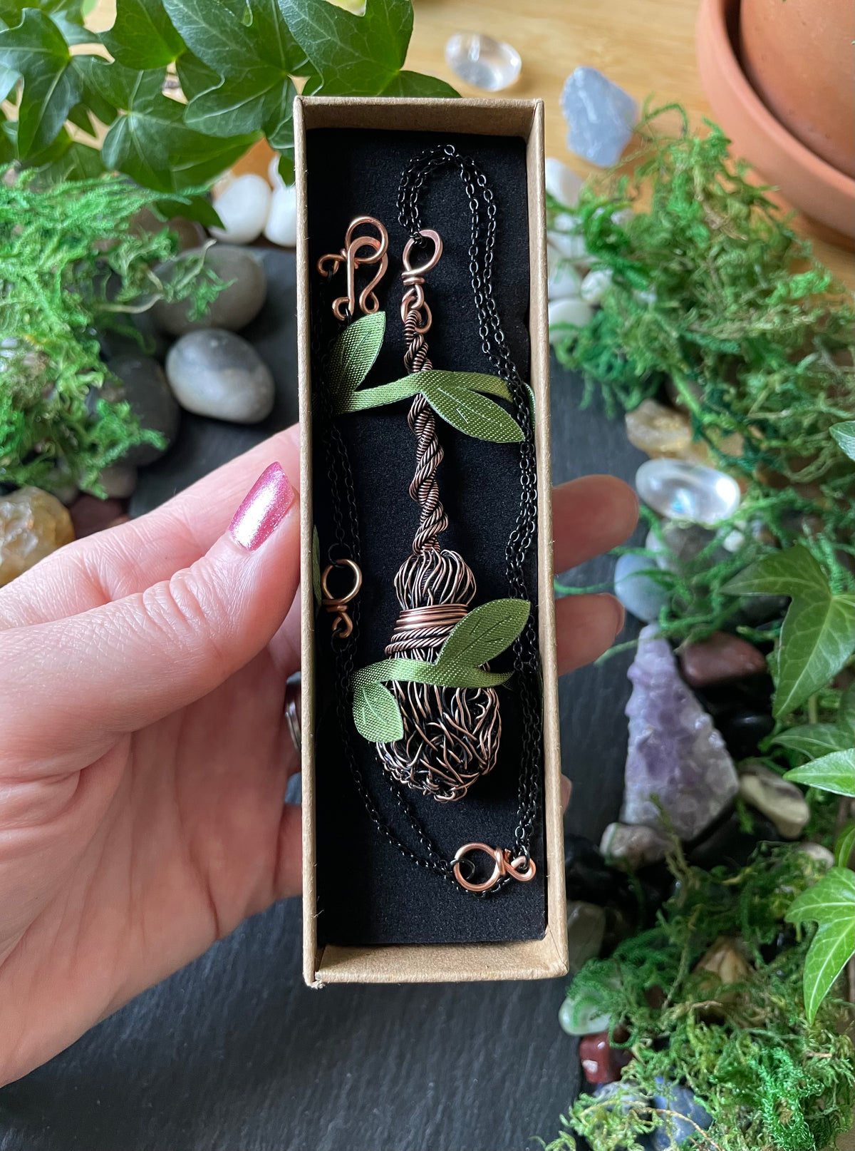 A hand holds a rectangular jewelry box containing a copper wire-wrapped pendant necklace with green leaf accents, surrounded by plants, stones, and moss.