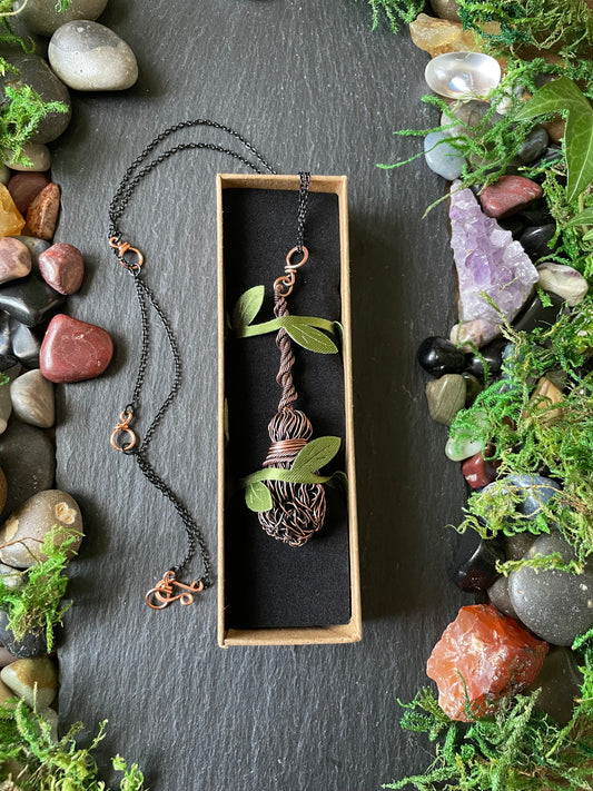 A handmade wire-wrapped pendant necklace displayed in a small box on a black background, surrounded by decorative stones and moss.