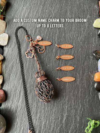A handmade copper wire broom pendant, displayed on a dark slate background with decorative stones. White text at the top reads 'ADD A CUSTOM NAME CHARM TO YOUR BROOM UP TO 8 LETTERS'.