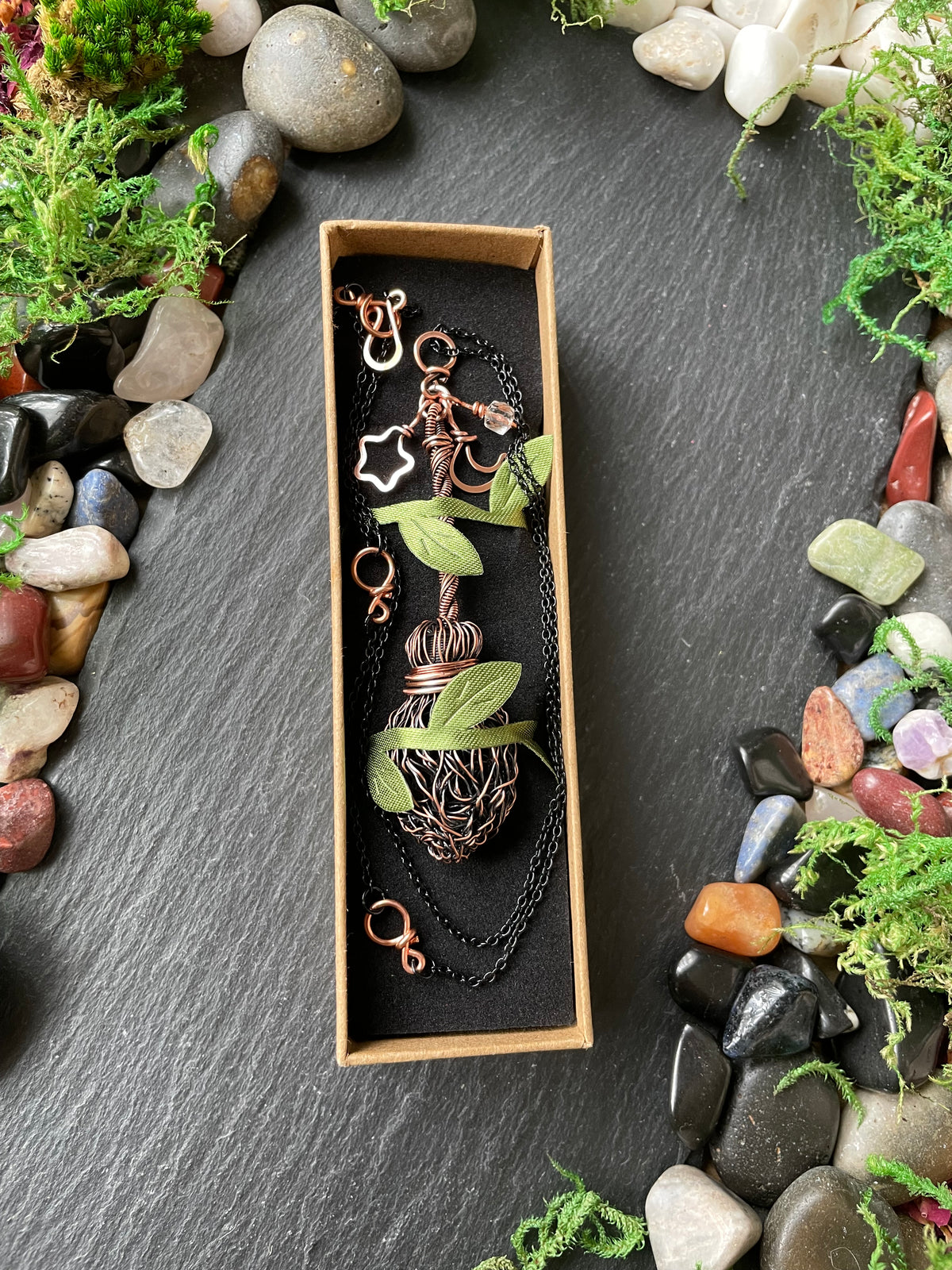 A mini metal handmade broom pendant, arranged on a dark slate surface surrounded by decorative stones and moss.