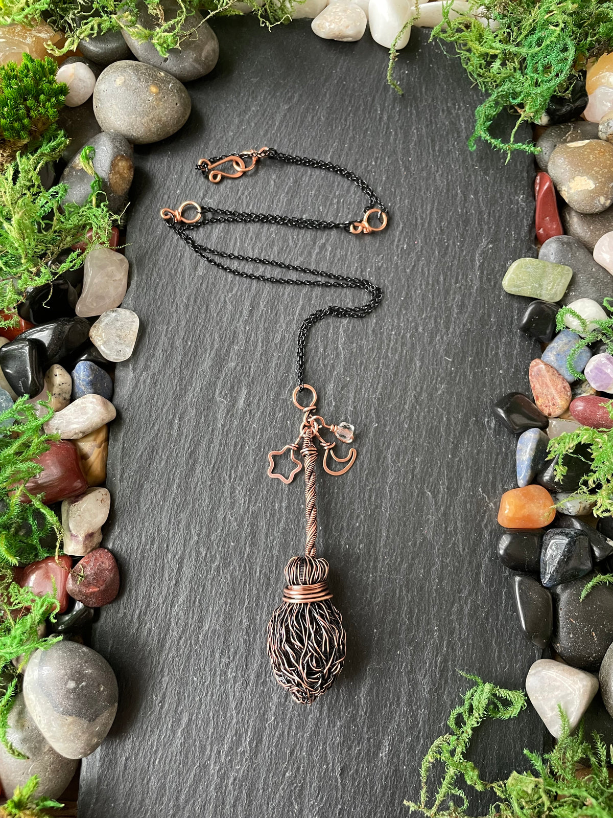 A handmade copper broom pendant necklace is displayed on a dark slate background, surrounded by a border of small stones and green moss. The pendant features a copper star and crescent moon charm. 