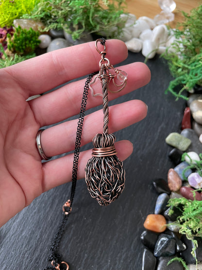 A handmade copper and black wire pendant necklace held in a person's hand, set against a dark slate background with decorative moss and stones.