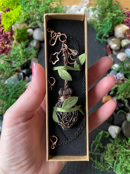 A hand holding a mini copper broom pendant, set against a background of green moss and small stones.