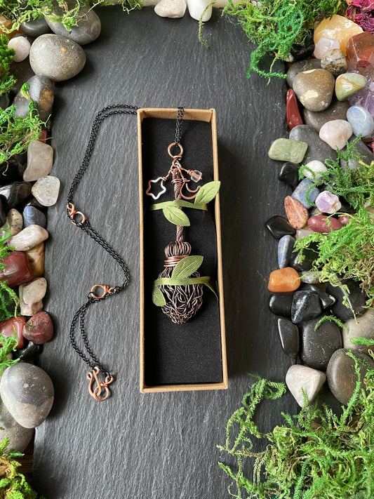 A handmade mini broom pendant, displayed on a dark slate background surrounded by stones and moss