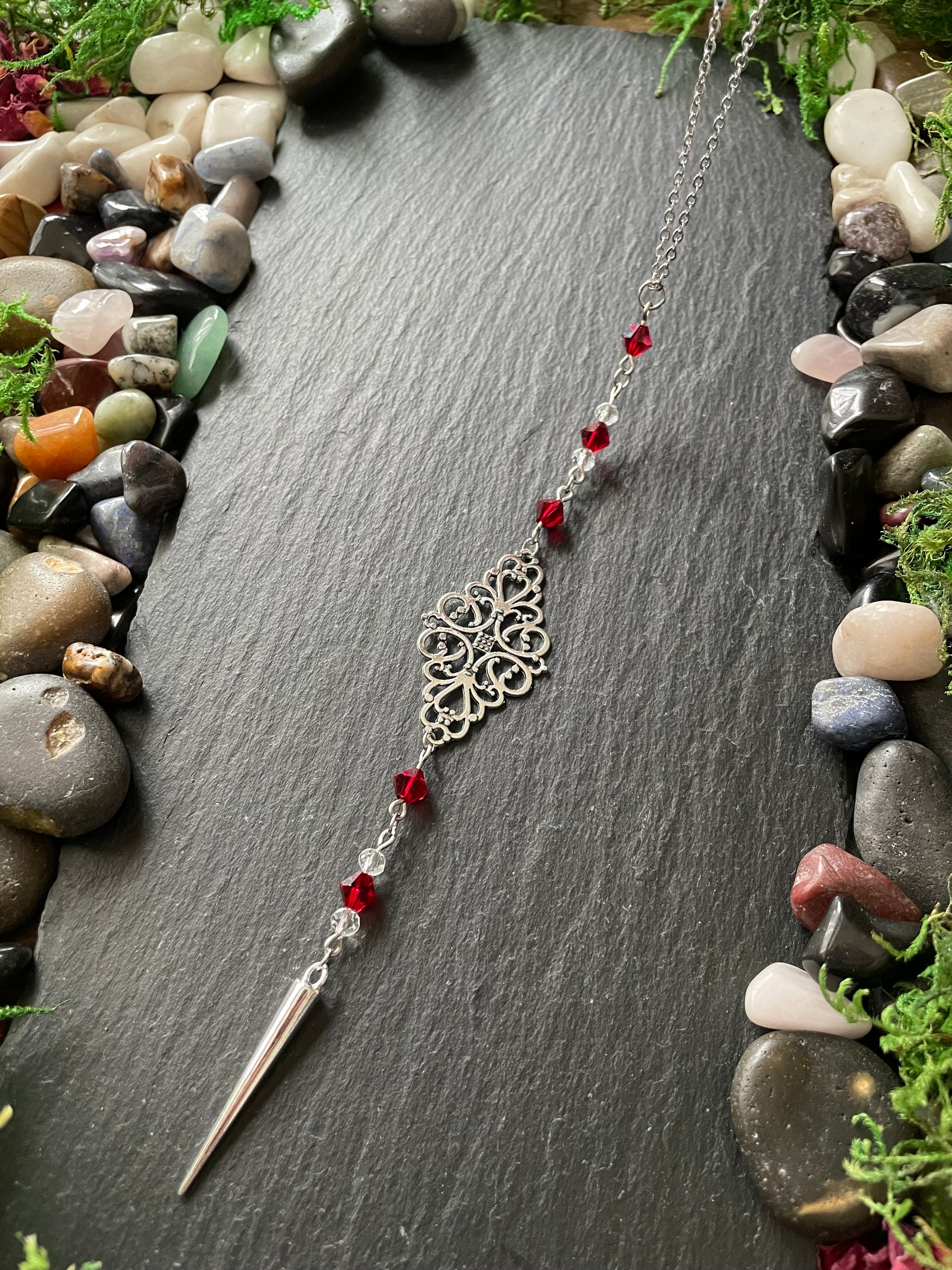 Silver car mirror charm with red beads on a dark stone surface surrounded by stones and greenery