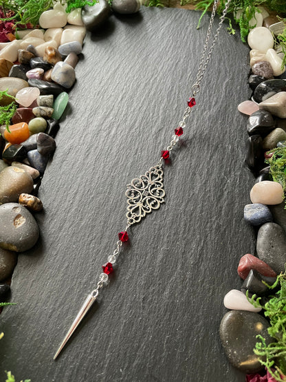 Silver car mirror charm with red beads on a dark stone surface surrounded by stones and greenery