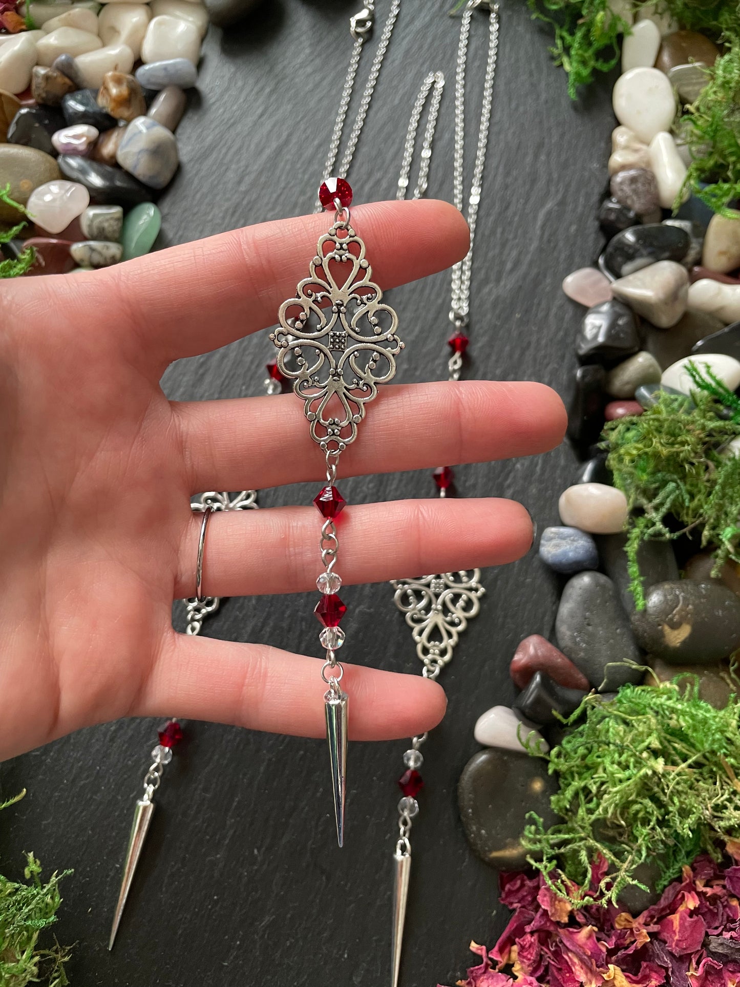 Hand holding a silver car mirror charm with red beads on a dark surface with decorative stones and plants.