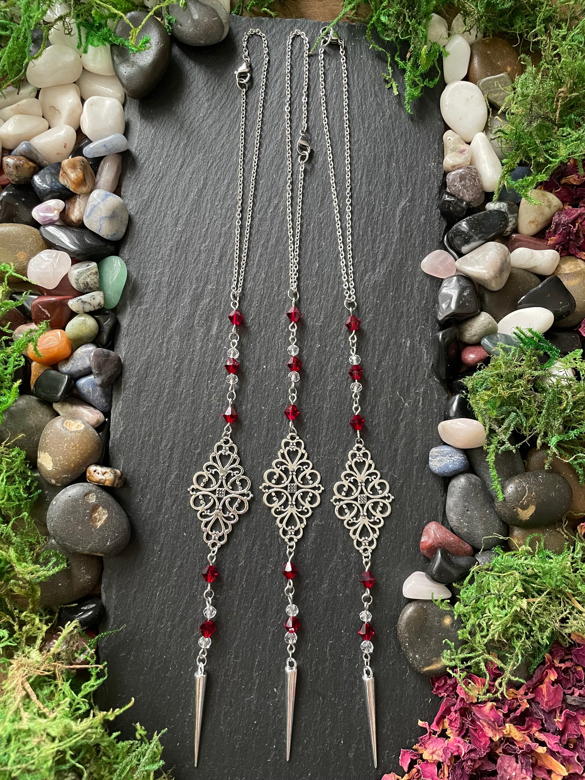 Silver car mirror charms with red beads on a dark slate surface surrounded by stones and plants