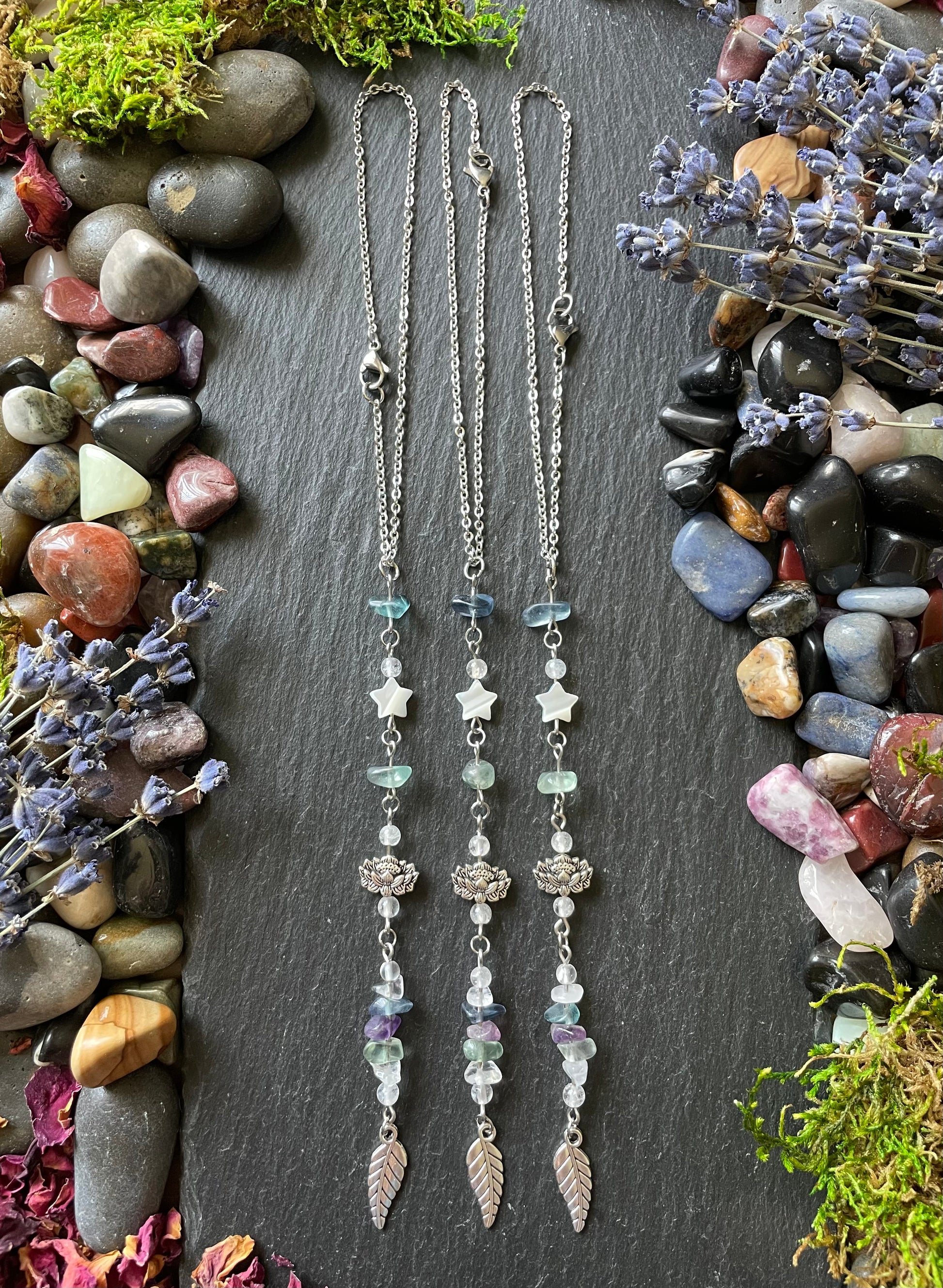 Three car charms with fluorite crystal beads, lotus flower beads, mother of pearl star beads, and leaf charms displayed on a stone slate surrounded by gems and moss