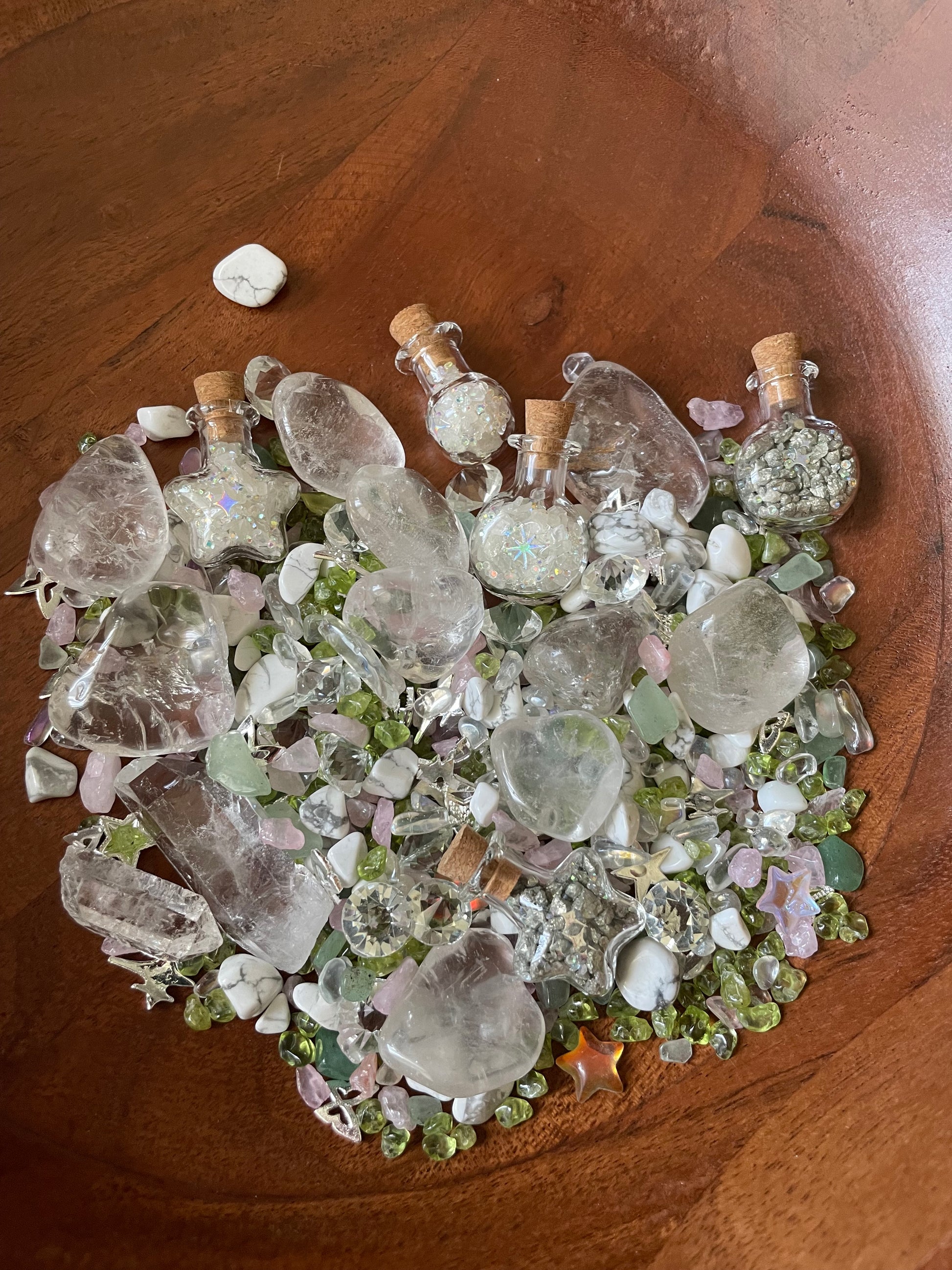 A pile of assorted clear quartz crystals, small glass vials with corks, and pale pink rose quartz chips mixed with green and white stones, arranged in a circular pattern on a dark brown wooden surface.