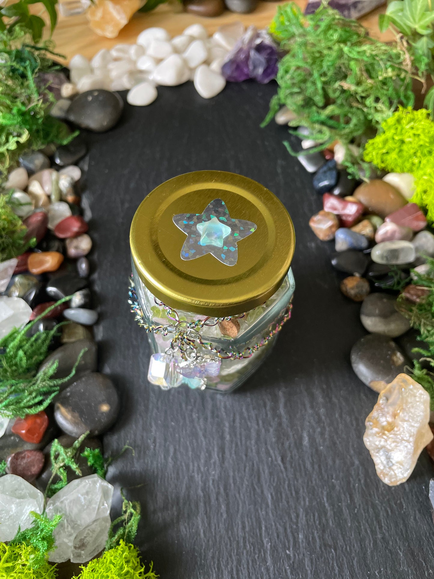 A small clear glass jar with a gold metal lid, surrounded by moss, stones, and crystals on a dark slate surface.