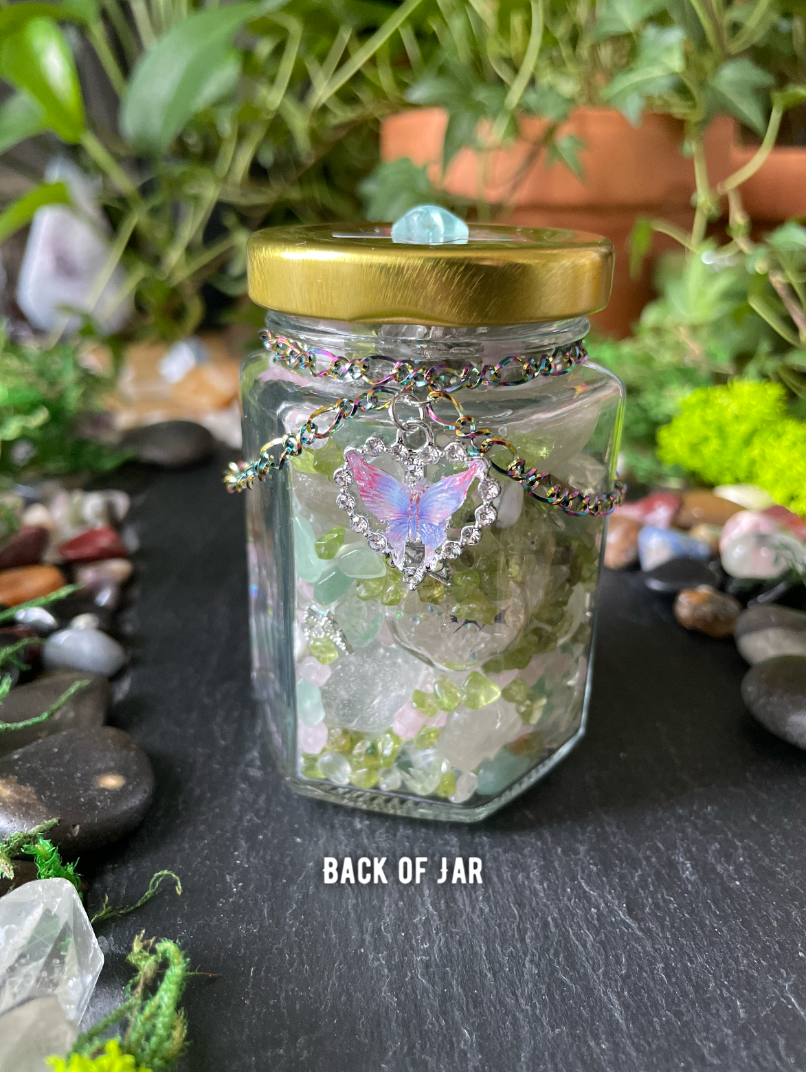 A clear glass jar with a gold metal lid, filled with white and green crystals, decorated with a silver chain and a heart-shaped butterfly charm. The jar is placed on a dark slate surface surrounded by green plants, moss, and decorative stones.