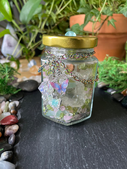 A small clear glass jar with a gold metal lid, filled with decorative elements including a butterfly-shaped charm, crystals, and small stones, photographed against a dark stone surface and green plants.