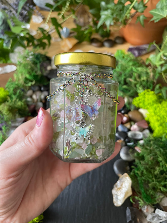 A hand holding a small clear glass jar filled with colorful crystals and butterflies, featuring a gold screw-on lid and a silver-toned chain.