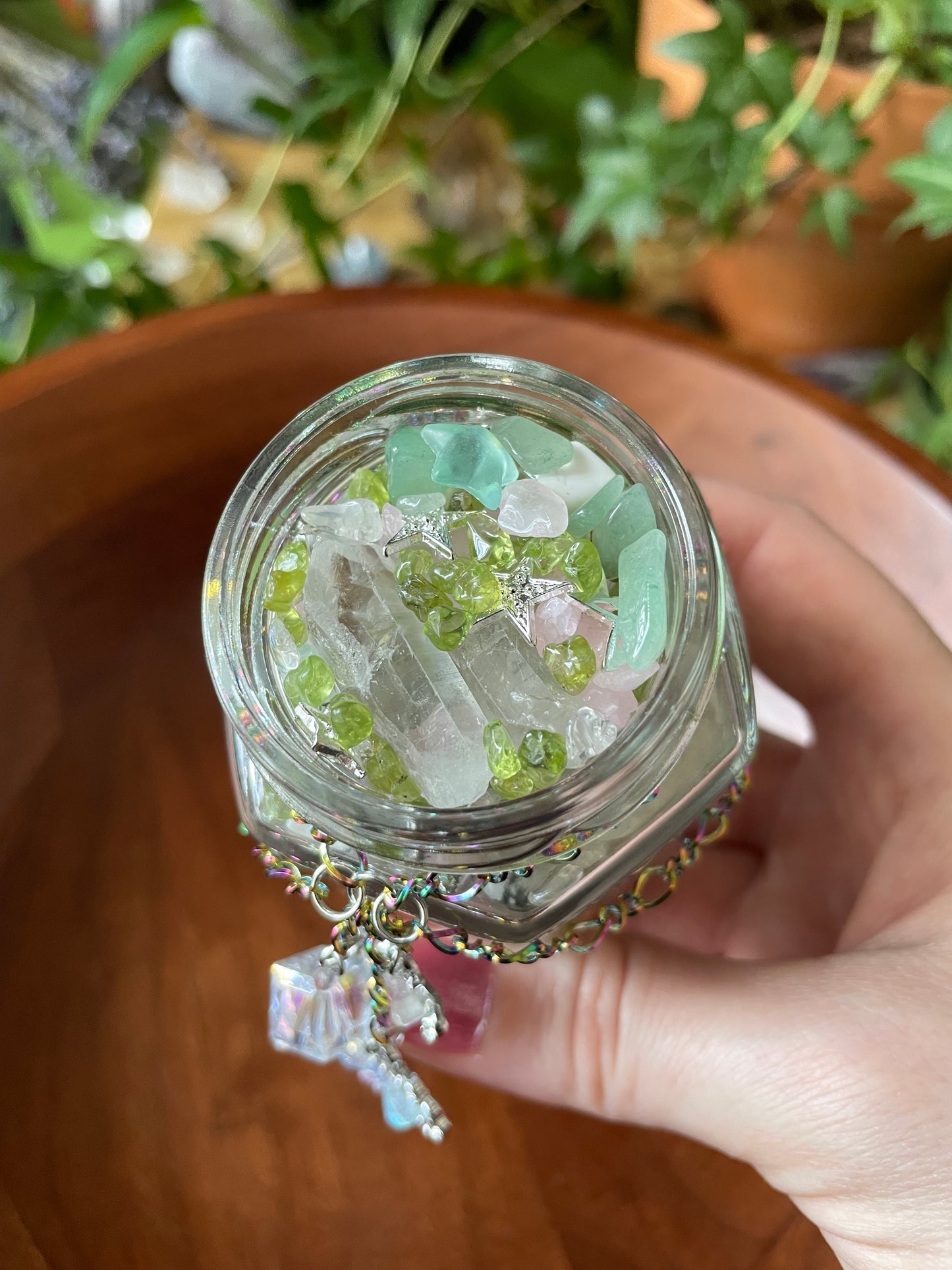 A hand holding a small clear glass jar filled with assorted crystals and gemstones, including pale green, white, and iridescent pieces, with a dangling charm, set against a warm brown wooden background.